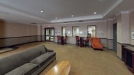 The Movie Lounge is a well-appointed, single-floor communal space designed to accommodate both relaxed socializing and private screenings. The open layout is anchored by a large projection screen mounted at one end of the room, complemented by a rich wood media cabinet with dark granite countertops beneath it. Recessed lighting illuminates the space throughout, casting a warm, even glow across the patterned carpet flooring that runs through the main areas.

Seating is arranged in several distinct configurations to encourage different types of interaction. A plush gray sofa and low wooden coffee table form a central lounge area, while clusters of high-top tables with red bar stools provide additional gathering spots. Cushioned armchairs in neutral tones with dark wood armrests are grouped near round side tables, creating comfortable conversational nooks. An orange wingback chair adds a pop of color near the perimeter.

Along one wall, a fully equipped kitchenette features dark cabinetry with granite countertops, a sink, stainless steel appliances including a refrigerator and dishwasher, and a microwave—supporting easy entertaining. Large windows along the exterior walls allow natural light to filter in, offering views of the surrounding brick building exterior. Framed artwork displayed throughout adds a refined, curated ambiance to the space.