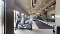 Located on Springside Lane in Middletown, CT, this single-floor fitness center offers a generously sized, open exercise space designed for a full-spectrum workout experience. The facility is defined by its striking vaulted ceiling with exposed industrial ductwork, recessed lighting, and sleek linear pendant fixtures that lend the space a modern, contemporary aesthetic. The interior is finished in a neutral gray palette throughout, with a combination of durable rubberized flooring and carpet workout zones that help delineate functional areas.

The cardio zone features a row of treadmills and elliptical machines positioned along large windows and glass doors, allowing natural light to pour in and offering views of the outdoor patio and surrounding grounds. Adjacent to this area, a functional training zone houses a multi-station cable and strength rig, a heavy punching bag, resistance bands, and a tire for dynamic conditioning exercises. A dedicated strength area includes a full dumbbell rack, selectorized weight machines, and benches for targeted muscle training.

Mirrored walls throughout the space enhance the sense of openness. Additional access doors on multiple sides provide convenient entry and exit, while ample windows ensure the environment remains bright and connected to the outdoors. The overall design balances functionality and modern aesthetics, making it a well-equipped fitness destination.