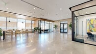 Located at 3170 W Olympic Blvd in Los Angeles, this single-floor commercial space presents a refined, contemporary aesthetic defined by clean lines and high-end finishes. Polished concrete floors extend throughout the interior, reflecting abundant natural light that pours in through expansive floor-to-ceiling windows overlooking the surrounding streetscape.

The layout flows openly from a welcoming entry corridor into a series of distinct functional zones. A bright lounge and seating area features marble-topped tulip tables paired with light wood and woven chairs, complemented by lush tropical plants that bring warmth to the modern palette. Large-format glazing frames unobstructed city views, enhancing the airy, light-filled atmosphere.

A spacious central hall showcases a prominent feature wall display, ideal for branding or presentations. Warm wood-paneled walls and ceilings appear throughout, providing a natural contrast to the crisp white surfaces. A dedicated lounge area with slatted wood ceiling details and large-format tile flooring opens toward a glass-enclosed stairwell enclosure, adding architectural depth.

Additional areas include a transitional entry vestibule with glass double doors and clean black-trimmed detailing, as well as a semi-outdoor lounge with slatted timber ceiling, generous seating, and views to the exterior. The space combines a professional, inviting atmosphere with flexible areas suited for leasing, retail, or community-oriented commercial use.