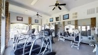 This fitness center offers a comprehensive workout environment designed to accommodate a range of exercise needs. The space is outfitted with an array of cardio equipment, including elliptical trainers and treadmills, alongside strength training machines, providing users with versatile options for their fitness routines. Large arched windows line one side of the room, flooding the interior with natural light and offering views of the surrounding greenery, creating an open and energizing atmosphere. Ceiling fans are strategically positioned throughout to maintain airflow and comfort during workouts. Full-length mirrored walls visually expand the space and allow users to monitor their form. Brick accent columns add a classic architectural detail, complementing the neutral tones of the carpeted flooring and white ceiling with exposed beam accents. Framed artwork is displayed on the walls, contributing to a polished and welcoming aesthetic. The layout is thoughtfully organized to maximize usable floor space while maintaining clear pathways between equipment, ensuring a safe and efficient experience for all users.