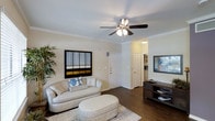 This single-story residence in Plano, Texas offers a thoughtfully designed living space with classic interior finishes. The living room serves as the centerpiece of the home, showcasing rich hardwood-style flooring that adds warmth and visual depth throughout. Crown molding lines the perimeter of the ceiling, lending an elegant, polished character to the space. A five-blade ceiling fan with an integrated light fixture is centrally positioned, providing both ambient lighting and efficient air circulation.

The neutral, warm-toned walls create a versatile backdrop, complementing a variety of furnishing styles. The layout flows naturally from the living area toward an entryway with a paneled front door, and a secondary door nearby suggests convenient access to additional rooms or storage. A corridor visible from the living space hints at further rooms beyond, including what appears to be additional living or utility areas. A media console area along one wall provides a practical and stylish anchor point. The overall ambiance is bright, clean, and inviting, reflecting a comfortable and functional single-level layout well-suited to everyday living in the Plano area.