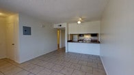 This single-floor space features an open-concept layout that connects the living area to the kitchen, creating a functional and cohesive environment. The living area is finished with large neutral-toned ceramic tile flooring, which extends throughout the main space and contributes to a clean, consistent aesthetic. A ceiling fan is centrally mounted, providing both lighting and ventilation. The textured popcorn ceiling adds a classic finish overhead, while white-painted walls keep the interior bright and neutral.

The kitchen is accessible via a pass-through counter with a dark laminate surface, which separates it from the living area while maintaining visual openness. Inside the kitchen, standard appliances are visible, including a white refrigerator and an electric range with a ventilation hood above. Dark upper cabinetry contrasts with the lighter surroundings. A hallway visible in the background leads to additional areas of the unit, suggesting a practical layout with private spaces set apart from the main living zone. An electrical panel is mounted on the living room wall near the hallway entrance. Overall, the space presents a straightforward and functional design.