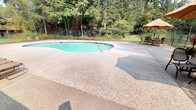 Located at 3418 SE Navigation Ln in Port Orchard, Washington, this outdoor pool facility offers a well-maintained recreational amenity set against a lush Pacific Northwest backdrop. The centerpiece is a generously sized freeform swimming pool with a kidney-shaped design, filled with clear blue-green water and equipped with dual stainless steel entry rails for easy access.

Surrounding the pool is a broad, textured aggregate concrete deck that provides ample space for lounging and circulation. A row of reclining lounge chairs lines one side of the pool, offering a dedicated area for relaxation. A patio umbrella and additional seating are positioned near the pool house, creating a shaded gathering spot.

The single-story pool house is clad in board-and-batten siding and features French doors with glass pane panels that open directly onto the pool deck. Posted signage at the entrance outlines pool rules and hours of operation, noting availability from 9:00 AM to 9:00 PM for residents and authorized guests. A first aid kit and emergency phone are mounted to the exterior wall, reflecting a commitment to safety.

The entire pool area is enclosed by a black iron fence with a gated entry, ensuring a secure environment. Towering evergreen trees frame the perimeter, providing natural privacy and a serene, forested ambiance characteristic of the greater Port Orchard region.