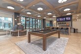 Located at 10 Executive Park Dr NE in Atlanta, Georgia, this single-floor amenity space blends sophisticated design with functional social areas. The lounge is defined by its striking coffered ceilings with warm wood beams, light herringbone-patterned flooring, and an abundance of natural light streaming through expansive floor-to-ceiling black-framed windows that open to a courtyard and pool area beyond.

The space is thoughtfully organized into distinct zones. A casual seating lounge features plush, patterned armchairs arranged around a gold-toned circular coffee table, positioned directly adjacent to the large sliding glass doors that lead to the outdoor terrace. A dedicated dining area offers rows of clean white tables with contemporary wood-and-gray chairs beneath a coffered ceiling with recessed lighting, framed by decorative lattice-style wood partitions that lend an open yet defined feel.

The entertainment area is a standout, anchored by a full-size billiards table set atop a neutral area rug, complemented by a curved banquette seating nook and wall-mounted screens above a bar counter. Artistic circular wall sculptures add a curated, gallery-like touch. A high-top work counter with bar-style seating rounds out the space, offering a versatile setting for both work and play within this well-appointed community hub.