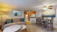 This single-floor unit is thoughtfully laid out to maximize functionality and comfort throughout. The entryway welcomes residents with tile flooring and a closet, opening into a combined living and dining area finished with wall-to-wall carpet and neutral gray walls. The living area is equipped with a ceiling fan and a window unit air conditioner, while a large window fitted with vertical blinds allows natural light to fill the space. A ceiling fan also serves the dining area, which comfortably accommodates a full dining set.

The kitchen is separated from the main living area by a half-wall pass-through with a wood-trimmed opening, maintaining visual connectivity while defining the space. It features warm honey-toned wood cabinetry, tile flooring, a double sink, a white refrigerator, and a white gas range with recessed lighting above.

A hallway off the main living area leads to the private rooms, including a carpeted bedroom with soft, neutral tones and access to a closet. A separate room functions well as a home office, featuring carpet flooring and ample wall space. The overall ambiance is calm and practical, with a cohesive neutral palette throughout.