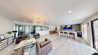 This thoughtfully designed single-floor community lounge offers a generous, open-plan layout that seamlessly blends functionality with contemporary style. Light-toned hardwood flooring runs throughout, lending warmth and continuity to the entire space.

The main living area is anchored by a striking branch-inspired chandelier and complemented by a second sculptural burst-style fixture, both serving as artistic focal points. Multiple distinct seating zones are furnished with rich brown leather sofas and armchairs, as well as a pair of vibrant cognac-leather accent chairs, creating intimate conversation areas within the open floor plan. Abstract artwork adorns the crisp white walls, adding visual character throughout.

A sleek, built-in bar counter runs along one side of the space, topped in white and framed by bold patterned tile backsplash in deep blue and geometric grey tones. Oversized gold dome pendant lights hang above the counter, which is lined with modern black barstools. A beverage station and under-counter refrigerator are also conveniently integrated nearby.

Large windows and glass-panel doors flood the space with natural light and provide views of an outdoor pool area. A steel-framed interior window wall separates an adjacent office or conference area while maintaining visual openness. The overall ambiance is modern, polished, and inviting.
