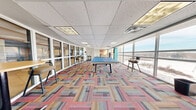 This energetic amenity space is designed to encourage socializing, recreation, and casual collaboration. The open floor plan is anchored by a full-size ping pong table and a foosball table, providing a lively focal point for the room. A vivid, multi-colored patchwork carpet in shades of coral, blue, tan, and gray adds a dynamic visual element underfoot, complementing the modern, playful aesthetic throughout.

One end of the space transitions into a lounge and meeting area, featuring a teal accent wall, a large whiteboard, and round high-top tables paired with contemporary bar-height seating — ideal for informal gatherings or casual work sessions. Additional high-top chairs are positioned near the windows for a more relaxed, scenic seating option.

Floor-to-ceiling glass windows line an entire wall, flooding the space with abundant natural light and offering sweeping views of the surrounding urban cityscape. Interior glass partitions provide visual connectivity to adjacent rooms while maintaining a sense of openness throughout.

The drop acoustic tile ceiling with recessed panel lighting ensures the space remains bright and well-illuminated. The overall design balances fun and functionality, making this a versatile destination suited for recreation, socializing, and collaborative activities alike.