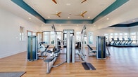 Located at 8925 Livingo Way in Kissimmee, Florida, this professionally designed fitness center spans a generous single-floor layout, offering a comprehensive and well-organized workout environment. The facility is divided into multiple functional zones, including a strength training area, a cardio zone, and a dedicated studio space, all connected by a central hallway.

The strength training area is outfitted with professional-grade Matrix cable machines, benches, and functional training rigs, set atop light hardwood flooring that runs throughout the facility. Tray ceilings with a bold teal accent band add architectural distinction, while multiple ceiling fans ensure comfortable air circulation. Large windows along the perimeter flood the space with natural light and offer views of the surrounding community.

The open studio room features full-length mirrors, TRX suspension trainers, a wall-mounted display screen, and a functional rack stocked with kettlebells and medicine balls — suitable for group classes or individual training. The cardio zone houses treadmills, ellipticals, and rowing machines near glass entry doors that open to the outdoors.

A support area with built-in cabinetry and a clean white countertop provides convenient storage and a front-of-house reception point. Recessed lighting and a clean, white-and-teal color palette give the entire facility a bright, modern, and welcoming ambiance.