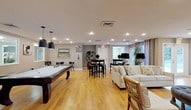 This well-appointed community lounge is a single-floor space designed to accommodate both recreation and relaxation. The open layout is anchored by warm-toned hardwood flooring that runs throughout, creating a cohesive and inviting atmosphere across all zones of the room.

At one end of the space, a full-sized pool table takes center stage beneath a pair of stylish matte black pendant lights, offering a dedicated area for recreation. Adjacent to it, framed wall art and decorative accents add a polished, finished look to the surrounding walls.

Moving through the room, a high-top bistro table with dark bar stools provides a casual dining or social gathering spot, while potted plants introduce a touch of greenery that softens the overall aesthetic.

The lounge area is furnished with a comfortable cream sofa and coordinating armchairs accented with patterned throw pillows, arranged to encourage conversation. Large French doors at the far end of the room flood the space with natural light and offer views of the outdoors, enhancing the open, airy feel. Recessed lighting throughout ensures the space remains well-lit at all times.