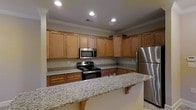 This single-floor space is designed with functionality and comfort in mind. The kitchen stands out as a central highlight, showcasing rich, warm-toned wood cabinetry that extends to the upper walls, providing generous storage throughout. The countertops are finished in speckled granite, offering both durability and a clean, classic aesthetic that complements the cabinetry. Stainless steel appliances, including a gas range, over-the-range microwave, and a full-size refrigerator, lend the kitchen a polished, modern feel. A peninsula with a corbel-supported granite overhang extends into the space, creating a natural breakfast bar or casual dining area that also helps define the kitchen zone. Recessed lighting and crown molding along the ceiling add refined architectural detail, enhancing the overall sense of quality. The neutral wall tones and light tile flooring keep the space feeling bright and open. The thoughtful layout of the kitchen allows for efficient workflow, with ample counter space between appliances and cabinetry. Overall, this space exudes a warm, inviting ambiance with timeless design choices suited for everyday living.