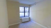 This single-floor space offers a clean, functional layout with a neutral and understated aesthetic throughout. The room features soft carpeted flooring that adds warmth underfoot, complemented by smooth, light-toned walls that create a bright and airy feel. A large window with dark-framed casings draws in natural light while offering views of the surrounding urban streetscape, giving the space a connection to the city outside. A white radiator beneath the window provides efficient in-unit heating, a classic fixture well-suited to urban residences. The white ceiling enhances the sense of openness, while simple white baseboards frame the room with a clean finish. The overall ambiance is minimal and move-in ready, with a blank canvas quality that lends itself well to a variety of interior styles and furnishing arrangements. A delicate hanging ornament near the ceiling adds a subtle decorative touch to the otherwise understated environment. The straightforward design of the space prioritizes practicality without sacrificing comfort, making it well-suited for those seeking a simple and efficient urban living arrangement.