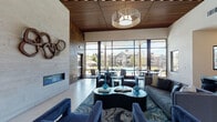 This contemporary clubhouse lounge is designed to impress, combining sophisticated materials and thoughtful layout to create an inviting community gathering space. The main lounge area commands attention with its soaring ceilings clad in warm slatted wood, which contrast beautifully against crisp white walls. A striking stone-clad accent wall serves as the focal point, anchored by a sleek linear electric fireplace and adorned with an eye-catching sculptural wood ring installation. Plush navy blue velvet seating, a bold organic-shaped coffee table, and a richly patterned area rug bring warmth and color to the space. Floor-to-ceiling window walls flood the interior with abundant natural light while offering unobstructed views of the outdoor pool courtyard and surrounding residential buildings, seamlessly blending indoor and outdoor living. A dedicated dining area with round tables and modern ghost chairs sits adjacent to the glazed facade, perfect for casual gatherings. The space also features a well-appointed amenity kitchen, complete with warm wood cabinetry, open floating shelves, a large island with bar seating, pendant lighting, stainless steel appliances, and a stone tile backsplash — ideal for entertaining. Throughout, the design balances contemporary elegance with functional comfort.