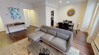 This single-floor residence offers a thoughtfully designed layout that balances comfort and functionality throughout. The open-plan living and dining area serves as the heart of the home, anchored by warm hardwood flooring and crown molding that lend a refined architectural character. Large windows flood the living room with natural light while offering views of the surrounding urban skyline, creating a bright and airy atmosphere. The living area accommodates a generous sectional sofa, a sleek coffee table, and a media console, while a decorative starburst mirror adds a stylish focal point above the dining table, which comfortably seats multiple guests. The galley-style kitchen is efficiently organized with rich dark cabinetry, white countertops, and full appliances, maximizing every inch of the space. Two well-proportioned bedrooms provide restful retreats; the primary bedroom features plush carpeting, crown molding, ample dresser storage, and access to a private en-suite area, while the secondary bedroom offers a similar serene aesthetic with soft carpet and a bright window. The bathroom is finished with clean white tile surrounding a full soaking tub and shower combination. Throughout the residence, a cohesive neutral palette and considered design details create a calm, inviting ambiance.
