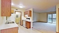 This single-floor unit presents an open-concept layout where the kitchen flows naturally into the adjacent living area, creating a cohesive and functional living space. The kitchen is equipped with medium-toned wood cabinetry offering ample upper and lower storage, white appliances including an electric range and refrigerator, and a light-colored countertop that extends into a peninsula-style breakfast bar — a practical feature for casual dining or additional prep space. The tile flooring in the kitchen provides a durable, easy-to-clean surface, while the living area transitions to carpeted flooring, adding a layer of comfort underfoot. A textured ceiling with a flush-mount light fixture illuminates the main living space. Visible through a doorway at the rear of the kitchen is a utility or storage area housing an electrical panel, suggesting thoughtful organization of mechanical elements. The neutral wall tones throughout contribute to a clean, understated aesthetic. The open passage between the kitchen and living room enhances the sense of space, making the layout well-suited for everyday living.