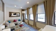 This 1-bedroom, 1-bathroom residence is designed with a clean, modern aesthetic that balances comfort and functionality across a single thoughtfully laid-out floor. The living area serves as the heart of the space, featuring large double-hung windows dressed with floor-length grommet curtains and adjustable blinds that offer excellent light control while framing views of the surrounding urban landscape. Recessed ceiling lighting provides consistent, even illumination throughout. The living room is finished with neutral-toned walls that complement a variety of décor styles, and the space is anchored by a bold, multicolored area rug that adds warmth and visual interest to the room. A wall-mounted television above a textured decorative frame adds a contemporary focal point to the space. The layout offers a natural flow that makes the most of the available square footage, ensuring each area feels purposeful and connected. The overall ambiance is modern and inviting, with an emphasis on natural light and urban connectivity. The combination of stylish finishes, generous window placement, and a neutral color palette creates a versatile and comfortable living environment well-suited for everyday living.