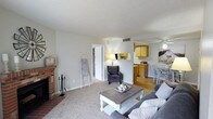 This single-floor residence offers a well-organized layout designed for comfortable everyday living. The living room serves as the heart of the home, anchored by a classic brick fireplace and complemented by plush carpeting and a sliding glass door that opens to a private patio, filling the space with natural light. A decorative wall clock and ladder shelf add character to the warm, neutral-toned interior.

Adjacent to the living area, the dining space connects seamlessly to the galley-style kitchen, which features warm wood cabinetry, light countertops, black appliances including a refrigerator and electric range, a stainless steel sink, and a skylight overhead that brightens the workspace.

The primary bedroom is generously proportioned, offering multiple windows, plush carpet flooring, a closet, and direct access to a private bathroom. The en-suite bath is clean and functional, featuring a single vanity and a shower-tub combination. A second bedroom accommodates twin beds comfortably, with shuttered windows that allow for natural light while maintaining privacy. A second full bathroom with wood-toned vanity cabinetry and a shower-tub combo serves this area of the home. Throughout, the space maintains a cohesive, neutral palette with textured ceilings and light, airy finishes.
