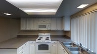 This two-floor residential space offers a practical and well-organized layout designed for comfortable everyday living. The interior is finished with warm, neutral wall tones that create a welcoming ambiance throughout.

The kitchen is a standout feature, equipped with ample white cabinetry that extends to the upper walls, providing generous storage space. Dark countertops offer a pleasing contrast against the bright white cabinets and appliances, which include a coil-burner electric range, an over-the-range microwave, and a double-basin sink. Overhead, a built-in fluorescent light fixture ensures the kitchen is well-lit. A window with vertical blinds allows for natural light to filter in.

The hallway connecting the kitchen to the living areas is clean and uncluttered, with a smooth, neutral-painted wall and a door that likely leads to a closet or utility space. The flooring transitions from dark hardwood in the kitchen to carpet in the adjacent living area, adding textural variety throughout the space.

The overall design is straightforward and functional, with a consistent color palette of whites, grays, and warm beiges tying the rooms together cohesively. The two-floor layout offers additional space and versatility for its occupants.