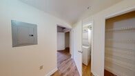 This single-floor loft-style unit blends industrial character with functional living in a compact yet efficient layout. The open main living area is the centerpiece of the space, showcasing dramatic exposed brick walls, heavy wood ceiling beams, and tall arched windows that flood the room with natural light. The hardwood floors run throughout, tying together the raw, industrial aesthetic with warmth and continuity.

The kitchen is positioned along one wall and features rich wood cabinetry, dark countertops, and a full suite of black appliances including a refrigerator, electric range, and dishwasher. An exposed brick accent wall and the soaring ceiling height above give the kitchen an open, airy feel that belies its compact footprint.

The hallway serves as a practical connector and utility hub, housing a stacked washer and dryer unit within a dedicated closet, along with an electrical panel and additional storage. The clean white walls of the hallway contrast with the industrial character of the main living space.

The bathroom is finished in a straightforward, clean style with small square tile flooring, a full soaking tub with shower, pedestal sink, and toilet — offering all essential amenities in a well-organized layout. Throughout the unit, the interplay of raw brick, natural wood, and white-painted surfaces creates a cohesive and distinctive loft ambiance.