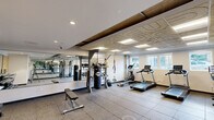 Located at 6300 9th Ave NE in Seattle's Roosevelt neighborhood, this fitness center offers a thoughtfully organized workout space across a single floor. The room is designed to maximize functionality, featuring durable rubber tile flooring throughout that provides both safety and comfort during exercise.

The cardio section is well-appointed with multiple treadmills, an elliptical trainer, and a stationary spin bike, all positioned near windows that allow natural light to filter in and offer views of the surrounding neighborhood. A multi-station cable machine anchors the strength training area, complemented by a full rack of dumbbells ranging across various weights, an adjustable workout bench, and a TKO heavy punching bag with medicine balls for combat-style training. Resistance bands and suspension training straps add further versatility.

Full-length mirrors line portions of the walls, visually expanding the space and allowing users to monitor their form during workouts. The ceiling features distinctive patterned acoustic tiles alongside recessed and panel lighting, ensuring bright, even illumination throughout. Exposed ductwork adds an industrial aesthetic detail to the space.

A water fountain is conveniently located near the entrance, and the room is accessed through a secure door. Overall, the fitness center delivers a comprehensive and practical workout environment suited for a wide range of exercise routines.