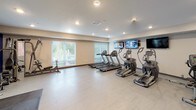 This fitness center is designed to support a comprehensive workout experience within a single, open floor plan. The space is divided into distinct functional zones, seamlessly integrated within a clean, modern layout. Light-toned flooring runs throughout, complemented by neutral walls and recessed lighting that keeps the environment bright and energizing.

The cardio area is well-stocked with multiple treadmills and elliptical machines positioned in rows, each with a clear sightline to a bank of wall-mounted flat-screen televisions. Large windows along one wall allow natural light to fill the space and provide views of the surrounding greenery, enhancing the workout atmosphere.

On the opposite end of the room, a bold royal blue accent wall anchors the strength training zone. This area features a full dumbbell rack with a flat utility bench, a multi-function training tower, and rubber mat flooring to accommodate floor exercises. A large wall mirror reflects the space, making it feel more expansive while allowing users to monitor their form.

Rubber exercise mats define dedicated workout areas, and the room includes two access doors for convenient entry and exit. The overall ambiance is functional, clean, and motivating, offering a well-rounded facility suitable for varied fitness routines.