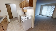 This single-floor unit presents a straightforward and functional layout, with the kitchen serving as a central feature of the space. The kitchen is equipped with a white electric coil-burner range and a matching full-sized refrigerator, providing all the essential cooking and storage necessities. Warm-toned oak cabinetry lines the upper walls, offering ample storage space, while a built-in range hood sits above the stove for ventilation. The kitchen floor features neutral tile, providing a practical and easy-to-clean surface. Adjacent to the kitchen, the living area transitions seamlessly via a subtle step down, where neutral wall-to-wall carpeting creates a softer, more comfortable atmosphere underfoot. Vertical blinds on the window allow for adjustable natural light, brightening the living space. The overall color palette is light and neutral throughout, contributing to a clean and open feel. The layout promotes easy movement between the kitchen and living areas, making the space well-suited for everyday living.