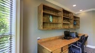 This single-floor space exudes a warm, rustic charm through its thoughtfully chosen furnishings and finishes. A standout feature is the built-in workstation, which showcases a richly textured wooden desk surface paired with wall-mounted open shelving crafted from reclaimed or distressed wood. The shelving units span a generous portion of the wall, offering ample display and storage space, and are accessorized with decorative pieces including a distinctive turquoise ceramic accent that adds a pop of color against the muted sage-green walls. Recessed lighting in the ceiling provides focused, clean illumination throughout the area, complementing the natural tones of the wood. Crown molding along the ceiling adds a subtle architectural refinement to the otherwise earthy, utilitarian design. The workspace is equipped with a desktop computer setup, making it a fully functional area for productivity. The combination of the soft wall tones, natural wood textures, and carefully placed décor creates a space that feels both organized and inviting — equally suited for focused work and relaxed browsing.