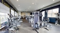 This fitness center offers a comprehensive workout environment designed to accommodate a variety of exercise routines. The space is laid out on a single floor, with an open floor plan that allows for easy navigation between equipment zones. Recessed ceiling lighting illuminates the entire area, complemented by natural light streaming in through windows along the perimeter walls, creating a bright and energizing atmosphere.

The cardio section lines one side of the room and features multiple treadmills, ellipticals, and stationary bikes, providing ample options for aerobic training. Toward the center and opposite side of the room, a robust selection of strength training machines is arranged, including cable systems and multi-function units such as the prominently placed Instinct machine. A dedicated free weights area rounds out the offerings, with a well-stocked rack of kettlebells and additional hand weights readily accessible.

The flooring consists of durable, rubber-style gym matting suited for heavy equipment and high-traffic use. Dark accent walls contrast with the lighter ceiling and trim, giving the space a modern, focused aesthetic. Ceiling fans aid in air circulation, and wall-mounted mirrors enhance the sense of openness throughout.
