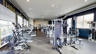 This fitness center offers a comprehensive workout environment designed to accommodate a variety of exercise routines. The space is laid out on a single floor, with an open floor plan that allows for easy navigation between equipment zones. Recessed ceiling lighting illuminates the entire area, complemented by natural light streaming in through windows along the perimeter walls, creating a bright and energizing atmosphere.

The cardio section lines one side of the room and features multiple treadmills, ellipticals, and stationary bikes, providing ample options for aerobic training. Toward the center and opposite side of the room, a robust selection of strength training machines is arranged, including cable systems and multi-function units such as the prominently placed Instinct machine. A dedicated free weights area rounds out the offerings, with a well-stocked rack of kettlebells and additional hand weights readily accessible.

The flooring consists of durable, rubber-style gym matting suited for heavy equipment and high-traffic use. Dark accent walls contrast with the lighter ceiling and trim, giving the space a modern, focused aesthetic. Ceiling fans aid in air circulation, and wall-mounted mirrors enhance the sense of openness throughout.