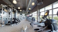 This fitness center offers a comprehensive workout environment designed for serious training and everyday exercise alike. The space is defined by its industrial-modern aesthetic, featuring exposed ductwork and piping along the dark ceiling, complemented by warm pendant lighting that adds an inviting ambiance to the otherwise functional setting. Floor-to-ceiling windows line one side of the facility, flooding the interior with abundant natural light and offering expansive outdoor views of surrounding greenery — an energizing backdrop for any workout. The cardio zone is prominently featured, with a long row of treadmills, elliptical machines, and WaterRower rowing machines arranged along the window wall for an open, airy feel. On the opposite side, a well-stocked strength and functional training area includes cable machines, weight systems, and specialty equipment such as an air bike, accommodating a wide range of fitness routines. The flooring is fitted with durable, cushioned carpet suited for high-traffic athletic use. A large mirror along the far wall enhances the sense of depth and space. Overall, the facility strikes a balance between professional-grade functionality and a modern, comfortable atmosphere.