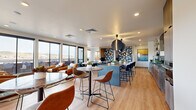 This contemporary lounge is a thoughtfully designed communal space that blends comfort, functionality, and modern aesthetics. Light hardwood flooring runs throughout, complementing the clean white ceilings fitted with recessed lighting. The space is organized into two distinct yet open areas that flow seamlessly together.

The social and dining zone features a large kitchen island with a dark matte countertop, surrounded by plush blue velvet bar stools and illuminated by striking oversized spherical pendant lights in matte black and brushed gold. Behind the island, stainless steel appliances, a hexagonal tile backsplash, open gold shelving with curated décor, and a wall-mounted flat-screen TV create a fully equipped and visually dynamic focal point. A geometric patterned accent wall adds an artistic layer to this area, alongside vibrant abstract artwork.

The lounge and seating area is anchored by warm cognac leather armchairs arranged on a flowing abstract area rug, paired with a concrete coffee table. A round marble-top dining table with upholstered charcoal chairs offers an additional gathering spot. Expansive black-framed floor-to-ceiling windows and sliding glass doors flood the interior with natural light while framing sweeping mountain views. The doors open directly onto an outdoor terrace, extending the social space further. The overall ambiance is inviting, sophisticated, and well-suited for both relaxed gatherings and productive socializing.
