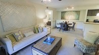 This single-floor residence features an open-concept layout that integrates the living and dining areas into one cohesive, functional space. The living area is anchored by a tufted light-gray sofa accented with decorative throw pillows in green and geometric patterns, complemented by a woven rattan coffee table that adds a natural texture to the room. A bold geometric wallpaper panel serves as a striking accent wall, lending visual interest to the otherwise neutral color palette of soft beiges and warm whites.

Overhead, a ceiling fan with a chandelier-style fixture provides both air circulation and ambient lighting, while track lighting above the dining area highlights a large abstract wall canvas in muted blues and greens. The dining space accommodates a round dark-wood table surrounded by upholstered chairs, creating an intimate setting ideal for small gatherings.

Plush wall-to-wall carpeting runs throughout the space, contributing to a warm and comfortable ambiance. A glimpse into an adjacent room reveals a bathroom or vanity area with dark wood cabinetry, indicating thoughtful storage solutions throughout. Overall, the space presents a clean, contemporary aesthetic with coordinated furnishings and carefully curated décor.
