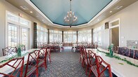 Located at 4 Stadium Dr in Angleton, TX, this single-floor gathering space offers a well-appointed layout designed for hosting events and group dining. The main dining area is the architectural centerpiece, showcasing a striking vaulted tray ceiling painted in a bold teal hue, accented by a sculptural multi-arm chandelier that adds warmth and elegance. Rows of white dining tables are paired with vivid red bamboo-style chairs featuring teal cushioned seats, creating a lively yet refined aesthetic. A richly patterned blue carpet covers much of the dining floor, complementing the colorful décor throughout.

Panoramic windows wrap around the sunroom-style dining area, flooding the space with abundant natural light and offering views of the surrounding landscaped grounds. Plaid drapery panels frame each window, adding a classic touch. Decorative teal wall panels and curated artwork contribute to the cohesive color palette.

The adjacent kitchen is equally well-equipped, featuring rich dark-stained cabinetry, light granite countertops, a central island, a mosaic tile backsplash, stainless steel appliances, and wood-look flooring. High windows bring in additional natural light. Together, these two areas form a versatile, thoughtfully designed space ideal for community gatherings, catered events, and group functions.
