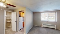 This single-floor unit offers a practical and comfortable layout designed for efficient everyday living. The main living area is finished with soft, neutral-toned carpet and painted walls in warm beige and gray hues, creating a calm and inviting atmosphere. A window allows natural light to filter into the space, and a baseboard radiator provides supplemental heating. The living area flows seamlessly into the kitchen through an open doorway, offering a connected feel between spaces.

The kitchen is thoughtfully equipped with warm honey-toned wood cabinetry both above and below the counter, offering generous storage. The countertops feature a light granite-style surface, complemented by a stainless steel sink and a full-size white refrigerator. The kitchen flooring transitions to a wood-look vinyl plank, adding a clean contrast to the carpeted living areas.

The bedroom is a generously sized room with plush carpet, neutral walls, and a ceiling fan with integrated lighting overhead. A closet and additional storage doors provide ample organizational space, and the room connects easily to the rest of the unit.

The bathroom is a full bath featuring a combination tub and shower, a toilet, and a vanity with sink. Wood-look vinyl plank flooring carries through here as well, and a window above the tub allows for natural ventilation and light.