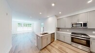 This well-designed 3-bedroom, 2-bathroom single-story home offers a clean, modern aesthetic defined by crisp white walls, light hardwood flooring, and an abundance of natural light. The open-concept kitchen and living area forms the heart of the home, where the kitchen stands out with gray shaker-style cabinetry, subway tile backsplash, quartz countertops, a full suite of stainless steel appliances including a gas range, built-in microwave, dishwasher, French door refrigerator, and a wine cooler. A center island with a sink adds both functionality and a natural gathering point. The living room benefits from expansive windows with views of lush greenery and direct access to an outdoor area via a glass-paned door, filling the space with natural light. Each bedroom features light hardwood flooring and windows that bring in daylight, with the primary bedroom offering generous proportions. Two closets provide ample storage, including a walk-in closet with built-in shelving and dual hanging rods. The two bathrooms are finished with light tile flooring and gray vanities; one includes a soaking tub and window, while the other features accessibility grab bars and a wall-mounted storage cabinet. Recessed lighting throughout enhances the home's clean, contemporary feel.