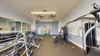This fitness center is a thoughtfully designed, single-floor workout facility that offers a comprehensive exercise environment for users of all fitness levels. The space is divided into distinct functional zones, seamlessly connected under a bright, open ceiling fitted with dual ceiling fans and suspended linear lighting that ensures consistent illumination throughout.

The strength training area is generously stocked with a full rack of dumbbells, multiple adjustable benches, a multi-station cable and weight machine, and a variety of free weights and accessories, including stability balls and medicine balls neatly organized on a dedicated rack. The cardio section, bathed in natural light from a wall of large multi-pane windows, houses treadmills, an elliptical, and stationary bikes, complemented by wall-mounted flat-screen TVs for entertainment during workouts.

Full-length mirrors line one wall, amplifying the sense of space and allowing for form checks during exercise. The rubber-floored surface provides both safety and comfort underfoot. A standout design feature is the bold motivational typography mural adorning the far wall, displaying words such as "Strong," "Focus," "Inspire," and "Empower" in vibrant orange and white lettering against a cool blue-grey backdrop. Entry is provided through a glass-paneled French door, with posted fitness center rules and a sanitization station ensuring a clean and organized environment.