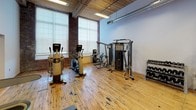 Located at 220 Merrimack in Lawrence, Massachusetts, this fitness center occupies a single-floor space that masterfully blends industrial heritage with modern athletic functionality. The facility retains the building's original character through warm hardwood floors, exposed brick walls, and rich wood-plank ceilings, all complemented by visible ductwork that adds to the loft-like aesthetic.

The main workout area is well-equipped with multiple treadmills, stationary bikes, and elliptical machines, alongside a multi-station cable and weight machine and a dumbbell rack. A wall-mounted flat-screen TV accompanies the cardio zone for an engaging workout experience. Large windows allow generous natural light to fill the strength and cardio areas, creating a bright and motivating atmosphere.

A dedicated climbing wall features colorful handholds and TRX suspension straps, paired with crash mats for safe training. Green turf flooring in this zone provides a functional surface for dynamic exercises. Vending machines near the entrance offer convenient refreshment access.

A glass-partitioned studio space with black steel framing separates an additional training or group exercise area, maintaining visual openness while defining distinct workout zones. The combination of industrial architecture and comprehensive fitness amenities makes this a well-rounded and visually compelling facility.