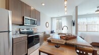 Located at 818 E Long St in Columbus, Ohio, this single-floor unit offers a thoughtfully designed open-concept layout that seamlessly connects the kitchen, dining, and living areas. The kitchen is well-appointed with rich dark-toned cabinetry, granite countertops, a full suite of stainless steel appliances including a refrigerator, electric range, and microwave, and a peninsula with pendant lighting that adds a stylish focal point. Light hardwood flooring flows throughout the space, enhancing the bright, airy atmosphere created by recessed lighting and large windows dressed with sheer curtains. The living area extends toward a sliding glass door that opens to a private balcony, inviting natural light and outdoor views. A tucked-away laundry closet houses a stacked washer and dryer for added convenience. The bedroom is a serene retreat with soft neutral tones and warm accent details. The bathroom features a granite-topped dark wood vanity with ample storage, a large mirror, and a glass-enclosed shower. The overall design balances modern finishes with a warm, functional aesthetic, making this unit an ideal urban residence in Columbus.