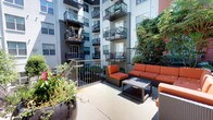 This outdoor courtyard amenity space is nestled within the interior of a modern multi-story residential building, offering a welcoming retreat surrounded by contemporary architecture. The space is thoughtfully designed with distinct zones for relaxation and socializing, connected by concrete pathways that guide residents through the area.

A generously sized patio seating area serves as a central gathering point, furnished with wicker sectional sofas and accent chairs fitted with bold orange cushions, arranged around a low coffee table. This arrangement invites casual lounging and conversation in an open-air setting flooded with natural sunlight. A separate, more intimate seating zone with darker wicker furniture provides a quieter alternative nearby.

Mature trees rise from raised concrete planters, casting dappled shade across the pathways and seating areas below. Additional landscaping includes lush shrubs, flowering plants, and ground-level greenery that soften the hardscape and add vibrant color throughout. Built-in concrete benches with wicker seat pads offer additional resting spots along the garden areas.

The courtyard is enclosed by the surrounding building's balconies, giving the space a sense of privacy while maintaining a bright, open atmosphere. Iron railings delineate level changes within the courtyard, creating gentle terracing that adds visual depth. The overall ambiance is one of urban tranquility, balancing modern design with natural elements.