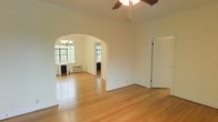 This single-floor space showcases a thoughtfully connected layout highlighted by a distinctive arched doorway that links two main living areas, adding a touch of classic architectural character. The warm-toned hardwood floors run continuously throughout, lending a cohesive and inviting feel to the interior.

The primary living area is equipped with a ceiling fan, providing both practical airflow and a casual lighting option. Through the arch, a second room is illuminated by a chandelier, adding a more refined lighting element to the space. Multiple windows in the adjoining room allow generous natural light to filter in, offering views of surrounding greenery and creating a bright, airy atmosphere.

Both rooms are finished with clean white walls that enhance the sense of openness and allow natural light to reflect throughout the space. A radiator and window-mounted air conditioning unit are visible in the far room, indicating year-round climate control. The overall ambiance is one of understated, classic charm — a versatile, well-lit space suited to a variety of uses and interior styles.