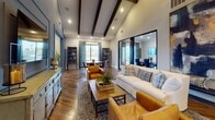 This thoughtfully designed single-floor community space blends modern sophistication with warm, inviting aesthetics. The open layout is anchored by striking herringbone-patterned hardwood flooring that flows throughout, while dramatic vaulted ceilings accented with dark exposed wooden beams lend an airy, elevated sense of volume to the main living area.

The lounge area serves as the heart of the space, furnished with a plush white sofa, leather accent chairs, and a woven coffee table adorned with lush greenery. Large-scale abstract artwork punctuates the white walls, adding bold visual interest. Adjacent to the lounge, a well-appointed kitchenette features white shaker cabinetry, dark subway tile backsplash, open floating shelves, a center island with bar seating, and a mounted television — ideal for casual gatherings.

A series of enclosed meeting or conference rooms branch off from the main area, each offering privacy with glass panel partitions and sliding barn-style doors. One room features a round wood dining table with leather chairs positioned beside sliding glass doors that invite natural light and outdoor views. Another intimate nook includes a neon accent light and mirrored wall detail, adding a contemporary, energetic touch. The overall ambiance balances professional functionality with residential warmth.