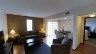 This single-floor residence offers a thoughtfully designed open-concept layout that seamlessly connects the living and dining areas, creating a cohesive and functional living environment. The living area is anchored by warm, dark hardwood flooring and features a comfortable seating arrangement centered around a sleek, dark coffee table. Earthy, neutral tones on the walls complement the space, lending it a cozy yet modern ambiance. A large area rug defines the seating zone, adding texture and warmth underfoot. Natural light flows generously into the space through multiple windows and a set of sliding glass doors fitted with vertical blinds, which lead directly to an outdoor area, blurring the boundary between interior and exterior living. The adjacent dining area is illuminated by recessed ceiling lighting and accommodates a dark wood dining table surrounded by a mix of seating. Ceiling vents are strategically placed throughout, ensuring efficient climate control. The overall design balances practicality with comfort, making it a welcoming and livable space suited for everyday use and casual entertaining.