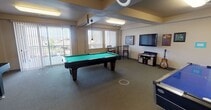 Located at 3304 E 44th Ave in Spokane, Washington, this single-floor community game room is designed for recreation and social interaction. The space is generously proportioned, with a layout that comfortably accommodates multiple gaming stations simultaneously. At its center sits a full-size pool table with a vibrant green felt surface, complemented by a long shuffleboard table along one wall and an air hockey table, providing a well-rounded selection of classic games. Overhead, flush-mounted fluorescent lighting illuminates the carpeted floor evenly throughout the room. A dedicated lounge nook features a high-top bistro table with bar-height chairs, positioned beside large vertical-blind windows that invite ample natural light and offer views of the surrounding exterior. On the opposite end of the room, a wall-mounted flat-screen television is flanked by framed artwork with an automotive theme, and a guitar on a stand adds a creative touch to the atmosphere. Additional décor, including plaques and small framed pieces, contributes to the room's personalized character. The space benefits from direct exterior access through a glass-panel door, further enhancing connectivity to the outdoors. Overall, the game room presents a welcoming, activity-rich environment suited for community gatherings and leisure.