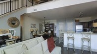 This two-story space makes a striking impression with its dramatic double-height ceilings and an open-concept layout that connects the main living areas with an airy, expansive feel. The main floor flows effortlessly from the living room to the dining area and kitchen, creating a cohesive and functional environment ideal for both everyday living and entertaining.

The living room anchors the space with a plush, light-toned sofa accented by rich, warm throw pillows, set atop dark hardwood flooring that runs throughout. The dining area features a substantial dark wood table surrounded by classic upholstered chairs, with an elegant chandelier overhead adding a refined focal point. A dark wood display cabinet nearby provides both storage and visual character.

The kitchen area is defined by a breakfast bar with cross-back bar stools and light stone countertops, offering casual seating alongside practical prep space. A bright white entry door is visible nearby, contributing to the bright, welcoming atmosphere.

Above, a second floor overlooks the main living area through a sleek metal railing, adding architectural depth and a loft-like quality to the interior. The interplay of the upper level, soaring ceilings, and open floor plan below creates a dynamic, multi-dimensional space that feels both grand and comfortably livable.