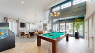 Located at 626 Shug Jordan Pkwy in Auburn, Alabama, this single-floor community clubhouse offers a thoughtfully designed gathering space that balances comfort, entertainment, and functionality. The open-concept layout connects the living and dining areas seamlessly, creating an inviting and versatile environment for residents.

The living area is a standout, featuring vaulted ceilings, light hardwood flooring, and an elegant gold chandelier that anchors the room. A wall-mounted flat-screen TV is flanked by built-in white cabinetry, while a mix of plush seating — including a rich amber leather sofa and sculptural accent chairs — gives the space a warm, modern aesthetic. An arcade game adds a playful touch to the lounge.

The dining and billiards zone transitions naturally from the living area, distinguished by a bold teal ceiling and large floor-to-ceiling grid windows that flood the space with natural light and offer views of the outdoor pool and greenery. A full-sized billiards table with a brass chandelier overhead completes the entertainment offering.

The dedicated office features an L-shaped marble-finish built-in desk, wood-look flooring, and a sleek wall sconce, providing a quiet and functional workspace. Throughout the clubhouse, the design blends contemporary finishes with warm, inviting details, making it an ideal community hub.