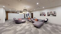 This thoughtfully designed single-floor space serves as a versatile lounge and collaborative work environment. The open layout is anchored by a striking geometric-patterned carpet in neutral gray tones, which lends visual depth and energy to the floor throughout. Two distinct seating zones are arranged within the main area — one featuring a large, modular gray sectional sofa accented with vibrant throw pillows and paired with sleek glass-and-chrome coffee tables, and another with a curved gray sofa and additional accent seating. Overhead, a bold, sculptural chandelier with an industrial-meets-artistic design serves as a standout focal point, complemented by recessed ceiling lighting that keeps the space bright and welcoming. A textured 3D white brick accent wall adds architectural interest to one end of the room, while warm wood-toned built-ins house a mounted flat-screen TV and integrated desk surfaces, providing functional workstation areas. A high-top table with bar stools offers a casual alternative for working or gathering. Decorative wall art and playful typographic signage infuse the space with personality. A secondary nook with two lounge chairs provides a quieter seating option near an interior corridor leading to adjacent rooms. The overall ambiance is modern, energetic, and professionally curated.