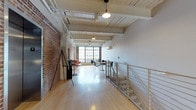 Roebling Lofts offers a single-floor open-concept loft space that masterfully blends industrial heritage with modern functionality. The space is defined by its soaring ceilings with white-painted wood planking and exposed structural beams, from which pendant light fixtures hang to provide warm, focused illumination throughout. Original exposed brick walls run along one side of the space, lending an authentic industrial character that contrasts beautifully with the smooth white walls on the opposite side. Polished hardwood floors in a light natural tone extend across the entire footprint, enhancing the brightness and openness of the interior. Large, multi-pane factory-style windows at the far end flood the space with generous natural light. A modern cable-railing staircase is positioned along one side, adding a contemporary architectural element. The open layout is currently arranged with seating and tables near the windows, making it a versatile setting suitable for a variety of uses. A wall-mounted split unit provides climate control, ensuring year-round comfort while maintaining the clean aesthetic of the space.