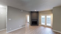 This single-floor Dallas property offers a well-designed layout with a clean, neutral aesthetic that creates a welcoming and functional living environment. The main living area is a standout, anchored by a charming corner brick fireplace that serves as a natural focal point. Light wood-style flooring runs throughout, complementing the soft gray and beige wall tones and lending the space a bright, airy feel. A textured ceiling adds subtle visual depth to the room. French doors with slatted blinds provide an elegant connection to the outdoors, inviting abundant natural light while maintaining privacy. Recessed lighting adds warm, directed illumination. Visible HVAC vents integrated into the walls and floors speak to the practical infrastructure of the home. An open passageway leads to an adjacent room, also filled with natural light from a large window, suggesting a seamless, open flow between living spaces. The overall ambiance is modern yet understated, offering a versatile canvas well-suited for a variety of interior styles. The combination of natural light, classic brick detailing, and cohesive finishes makes this Dallas property an inviting and functional place to call home.