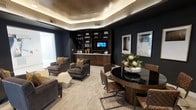 This refined single-floor space exudes a contemporary, upscale ambiance defined by deep charcoal walls, warm wood accents, and thoughtfully curated furnishings. A striking coffered ceiling with integrated ambient uplighting creates a soft, golden glow that highlights the textured ceiling finish, lending the room a sense of intimate sophistication. The built-in shelving and media unit along the back wall provides both functional storage and a polished focal point, complete with a mounted flat-screen television and tastefully arranged décor. The lounge area features plush, high-backed armchairs upholstered in rich textured fabric, arranged to encourage conversation and relaxation. A geometric stone side table adds an artistic accent to the seating arrangement. Complementing the lounge, a round dark-topped dining or meeting table is surrounded by quilted leather chairs with sleek metal frames, making the space equally suited for social gatherings or professional use. Natural light filters softly through frosted window panels, balancing the moody interior palette. The neutral toned carpet grounds the space, while a fresh floral centerpiece introduces an organic contrast to the room's dark, luxurious aesthetic. Overall, this space blends elegance with functionality in a polished, contemporary design.