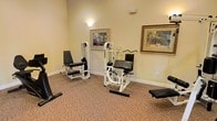 This single-floor fitness space is designed to provide a functional yet welcoming environment for strength training and exercise. The room features wall-to-wall carpeting in a warm, neutral tone that adds a layer of comfort underfoot, distinguishing it from a typical gym setting. Multiple white-framed resistance and strength-training machines are thoughtfully arranged throughout the space, including leg extension and multi-function cable machines, offering a range of workout options. A padded exercise bench is also present, adding versatility to the available equipment. Soft, ambient wall sconces cast a warm glow throughout the room, complementing the natural light and creating an inviting atmosphere. The walls are painted in a soft cream tone and adorned with framed artwork featuring tropical and outdoor scenes, contributing to a relaxed, resort-like ambiance. White crown molding and baseboards provide clean architectural detail throughout. The overall design balances practicality with aesthetic comfort, making this fitness room a pleasant space for residents or guests to engage in their exercise routines.