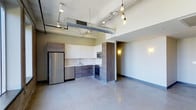 This Dallas residence showcases a striking industrial-modern aesthetic across a single open-concept floor. The space immediately impresses with its soaring ceilings, exposed ductwork, and polished concrete floors that lend an authentic urban loft character throughout. Edison-style pendant lighting hangs from above, adding warmth and a stylish industrial touch to the open living area.

The kitchen is thoughtfully designed with a clean, contemporary palette, featuring a combination of white upper cabinetry and wood-toned lower cabinets. Stainless steel appliances — including a full-size refrigerator, built-in microwave, and range — provide a sleek, functional workspace. The countertops and under-cabinet sink complete the kitchen's modern, efficient layout.

The open living area flows seamlessly from the kitchen, offering a generous, flexible space bathed in ambient light from wall sconces and pendant fixtures. The polished concrete flooring ties the entire space together, reinforcing the industrial design language while remaining low-maintenance and durable. The minimalist wall finishes keep the aesthetic clean and versatile, allowing for a wide range of interior design approaches. Overall, this space delivers a compelling blend of modern functionality and urban industrial character.